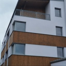 SKLADOKIEN.PL Sp. z o.o. - Nowoczesny budynek z szarymi oknami i drewnianą elewacją, widziany pod kątem z dołu, z balkonem na górze i pochmurnym niebem w tle.