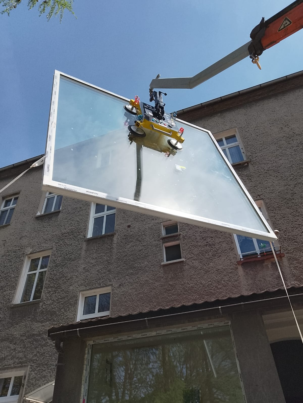 Montaż dużego okna z białym profilem za pomocą przyssawki na ramieniu dźwigu, na tle szarej elewacji budynku z oknami.