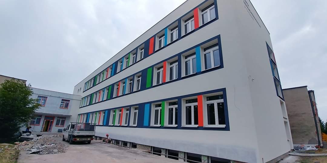 Budynek szkoły po renowacji elewacji z nowymi oknami i kolorowymi elementami dekoracyjnymi. Widoczny fragment terenu budowy i samochód dostawczy.