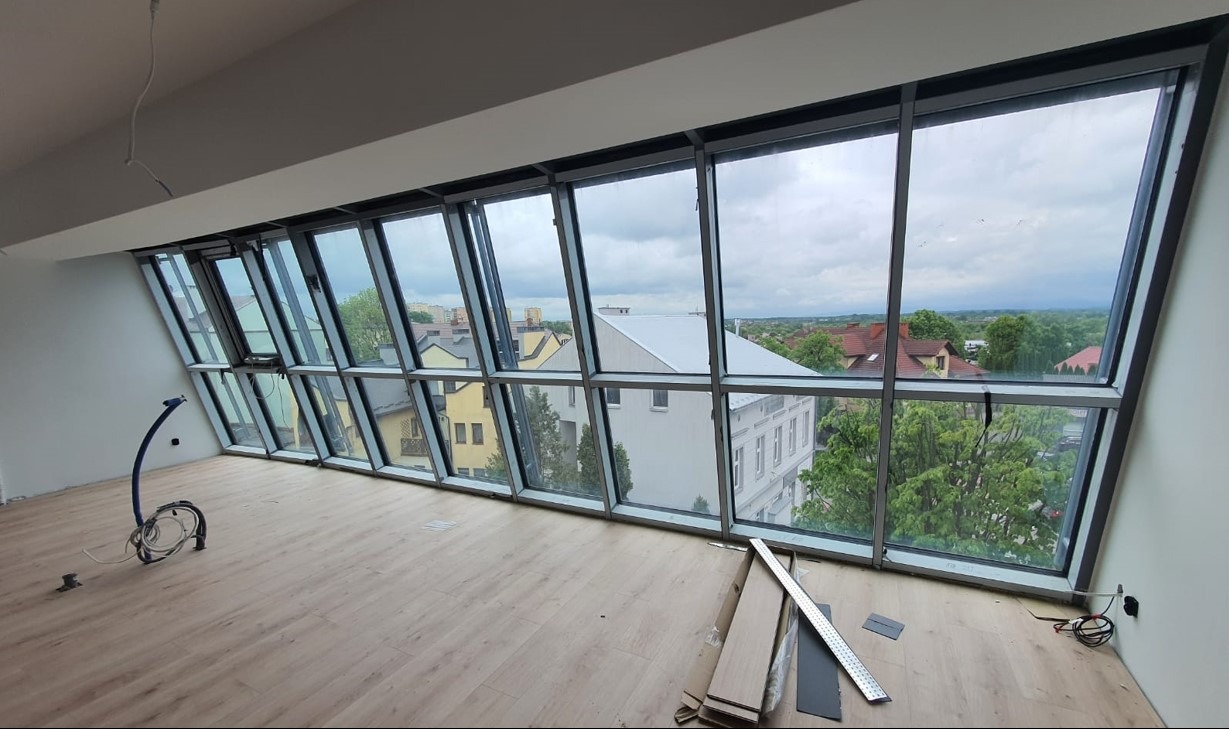 Nowoczesne okna z aluminiową ramą, skośne, z widokiem na miasto. Wnętrze w trakcie wykańczania, widoczne narzędzia i materiały budowlane.