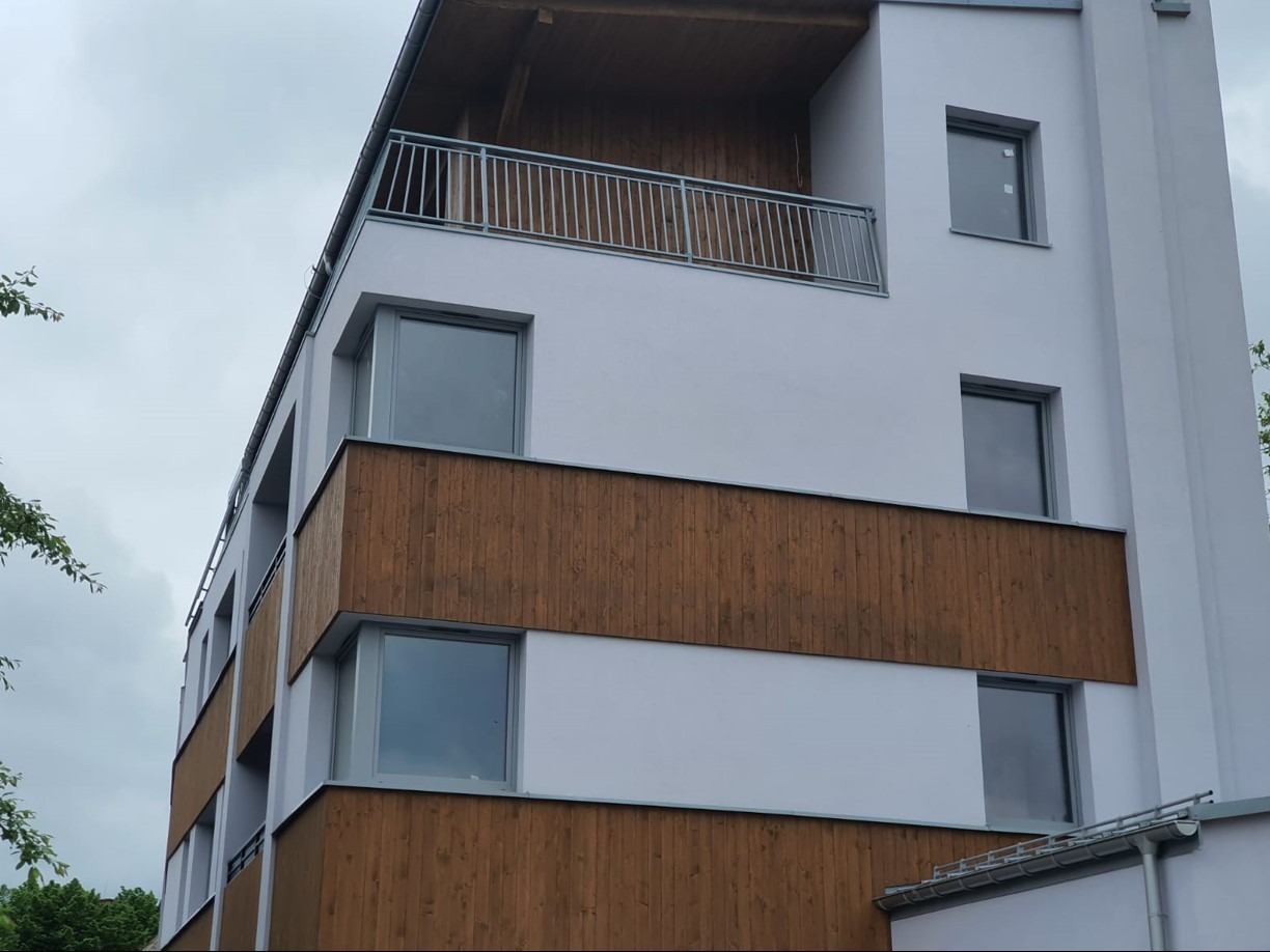 Nowoczesny budynek z szarymi oknami i drewnianą elewacją, widziany pod kątem z dołu, z balkonem na górze i pochmurnym niebem w tle.