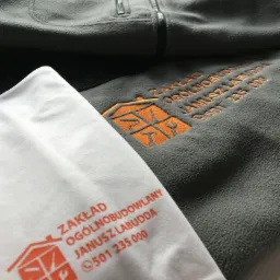 Szara bluza polarowa z pomarańczowym haftem logo firmy budowlanej oraz biała koszulka z nadrukowanym tym samym logo.