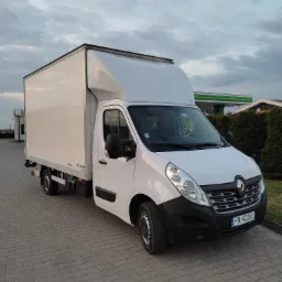Biały samochód dostawczy Renault Master z zabudową kontenerową na parkingu z kostki brukowej, widok z przodu, w tle stacja benzynowa i zachmurzone niebo.