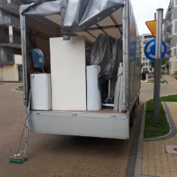 Otwarta paka samochodu dostawczego wypełniona meblami i sprzętami AGD zabezpieczonymi folią, drabina oparta o bok pojazdu, na chodniku leży miotła.