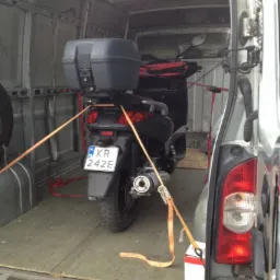 Skuter zabezpieczony pasami wewnątrz furgonetki, gotowy do transportu. Widoczne tablice rejestracyjne i elementy mocujące ładunek.