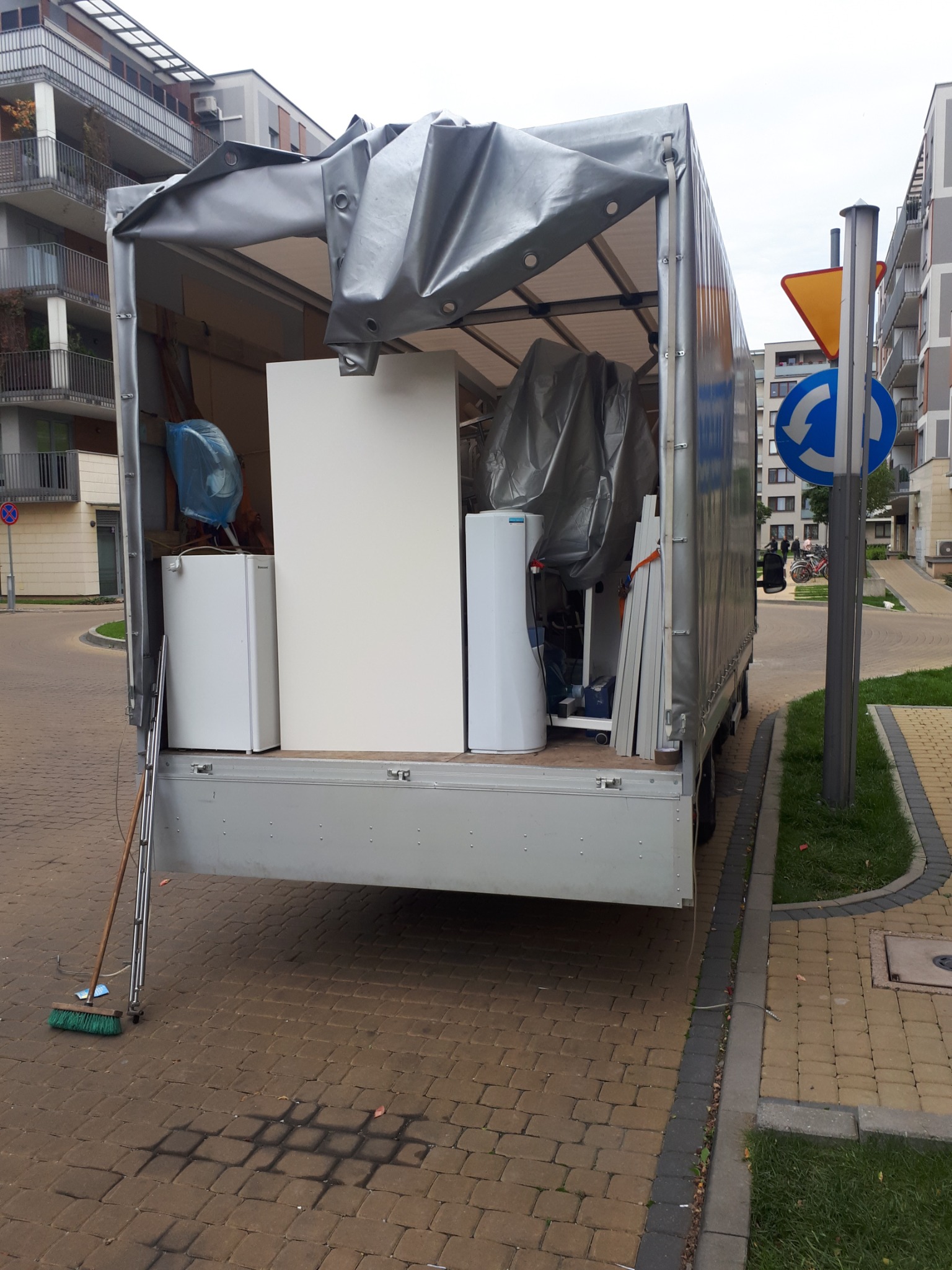 Otwarta paka samochodu dostawczego wypełniona meblami i sprzętami AGD zabezpieczonymi folią, drabina oparta o bok pojazdu, na chodniku leży miotła.