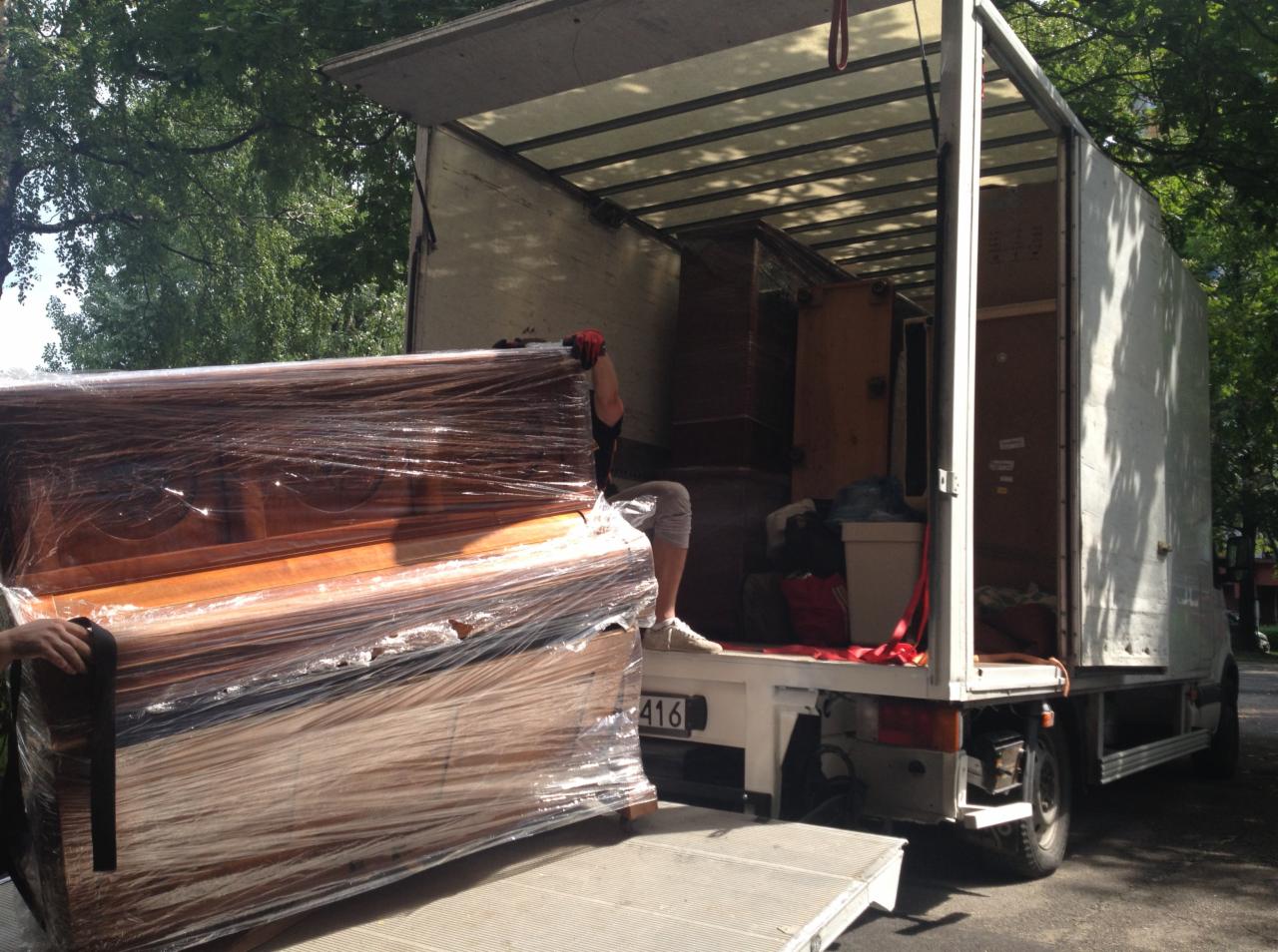 Pianino owinięte folią na rampie załadunkowej busa. Widoczne meble wewnątrz pojazdu i osoba zabezpieczająca ładunek. Usługa transportowa w trakcie realizacji.