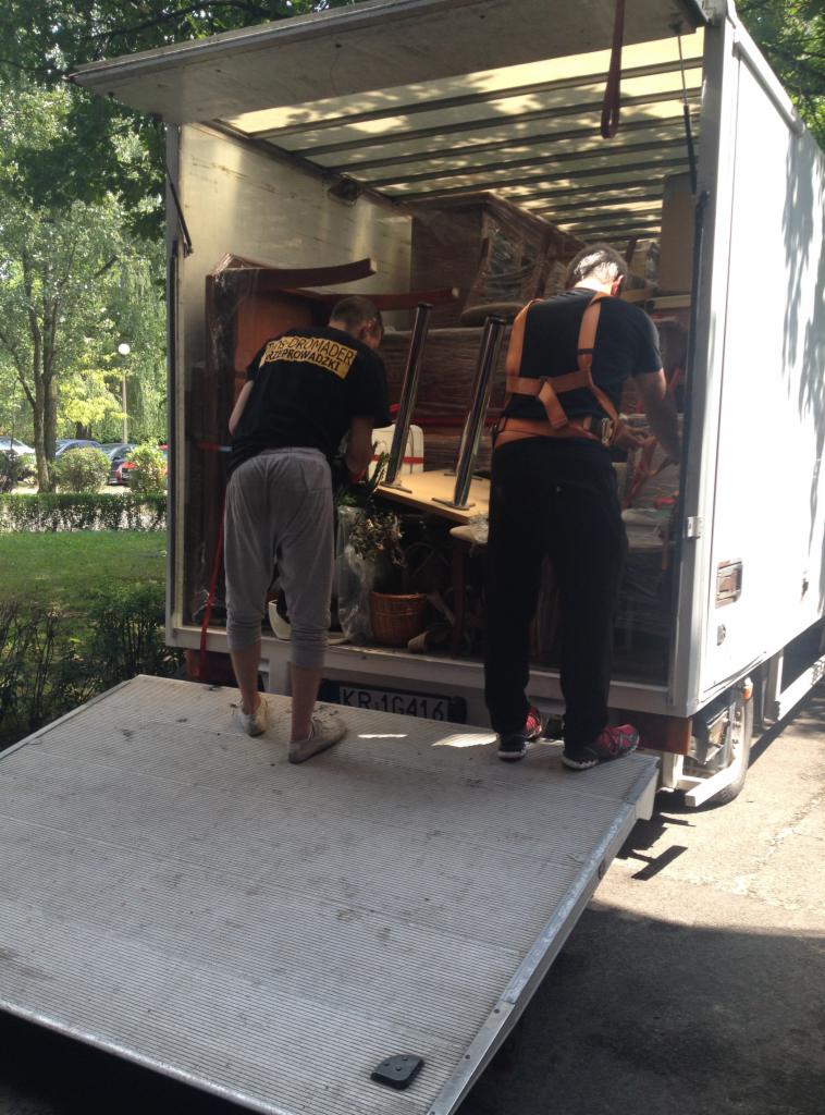 Pracownicy w trakcie załadunku mebli do białego busa z otwartą rampą, widoczne drzewa w tle. Jeden z nich w czarnej koszulce z napisem.