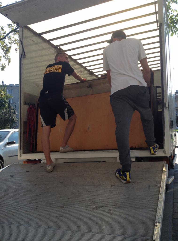 Pracownicy w trakcie załadunku mebla do busa z rampą. Jeden z nich ma koszulkę z napisem 'Romader Przeprowadzki'.