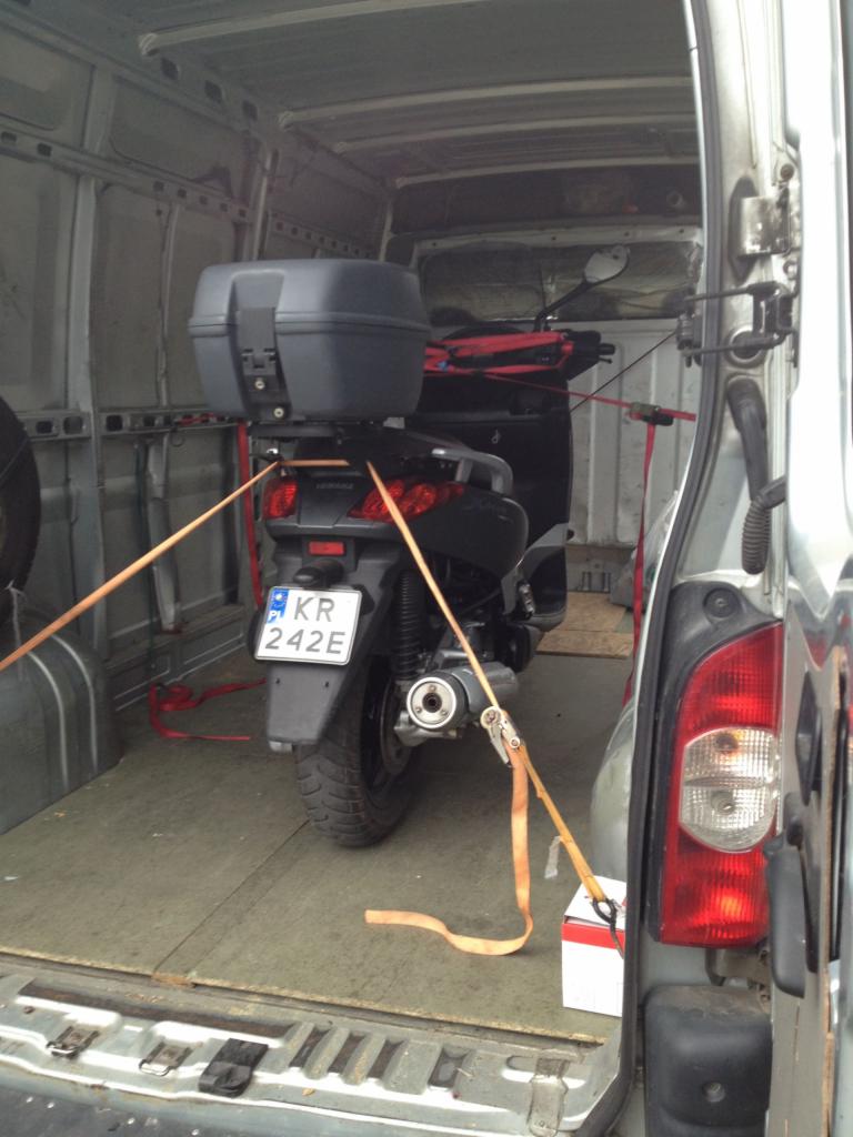 Skuter zabezpieczony pasami wewnątrz furgonetki, gotowy do transportu. Widoczne tablice rejestracyjne i elementy mocujące ładunek.