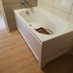 Nowa, biała wanna z obudową panelową ustawiona w rogu łazienki, z widoczną baterią i słuchawką prysznicową w kartonie wewnątrz, podłączaną do instalacji.