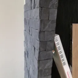 Fragment ściany wykończonej szarą, kamienną okładziną dekoracyjną, z widocznym narożnikiem i opakowaniem paneli podłogowych Kronopol w tle.