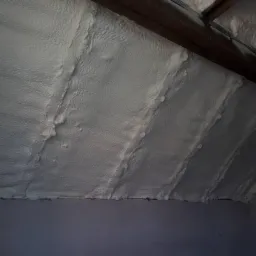 Poddasze ocieplone białą pianką natryskową, widoczne drewniane elementy konstrukcji dachu i szara ściana, nierównomierna struktura pianki.