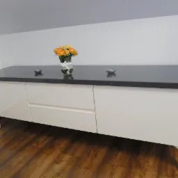 Biała komoda na giętych nóżkach z szarym blatem, ozdobiona kwiatami i świecznikami, na tle skośnego sufitu i drewnianej podłogi.