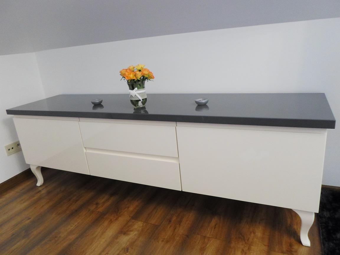 Biała komoda na giętych nóżkach z szarym blatem, ozdobiona kwiatami i świecznikami, na tle skośnego sufitu i drewnianej podłogi.