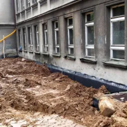 Odsłonięte fundamenty budynku z widoczną czarną folią izolacyjną i żółtą rurą drenażową, otoczone ziemią. Prace ziemne w toku.
