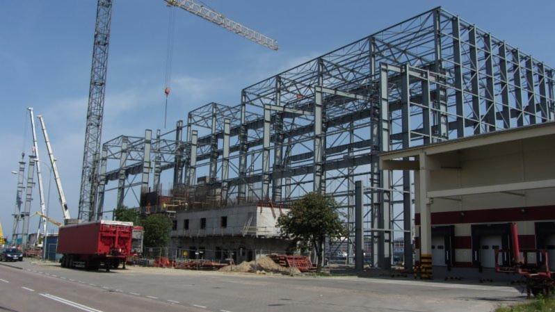 Wznoszenie stalowej konstrukcji budynku przemysłowego z widocznymi elementami betonowymi, żurawiami wieżowymi i transportem ciężarowym w tle.