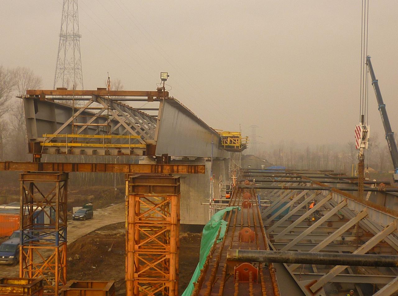 Montaż przęsła mostu stalowego w trakcie budowy, widoczne elementy konstrukcyjne, dźwig, oraz podpierające stalowe rusztowania w mglisty dzień.