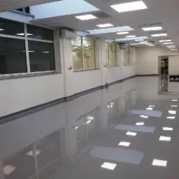 Długi korytarz z połyskującą, szarą podłogą żywiczną, odbijającą światła sufitowe i okna. Białe ściany z szarą listwą przypodłogową. W tle widoczne otwarte drzwi.