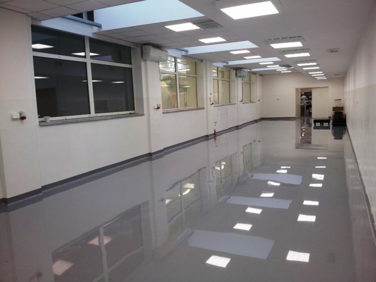 Długi korytarz z połyskującą, szarą podłogą żywiczną, odbijającą światła sufitowe i okna. Białe ściany z szarą listwą przypodłogową. W tle widoczne otwarte drzwi.