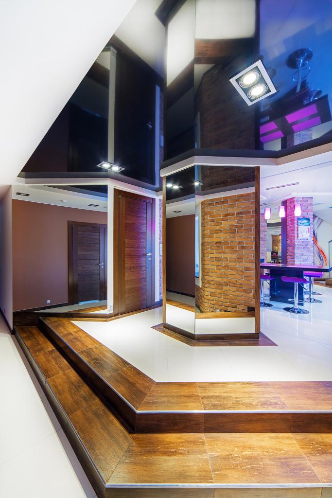 Nowoczesne wnętrze z lustrzaną kolumną obłożoną cegłą, ciemnym sufitem napinanym i schodami wyłożonymi płytkami imitującymi drewno. Widoczne drzwi i bar z fioletowym oświetleniem.