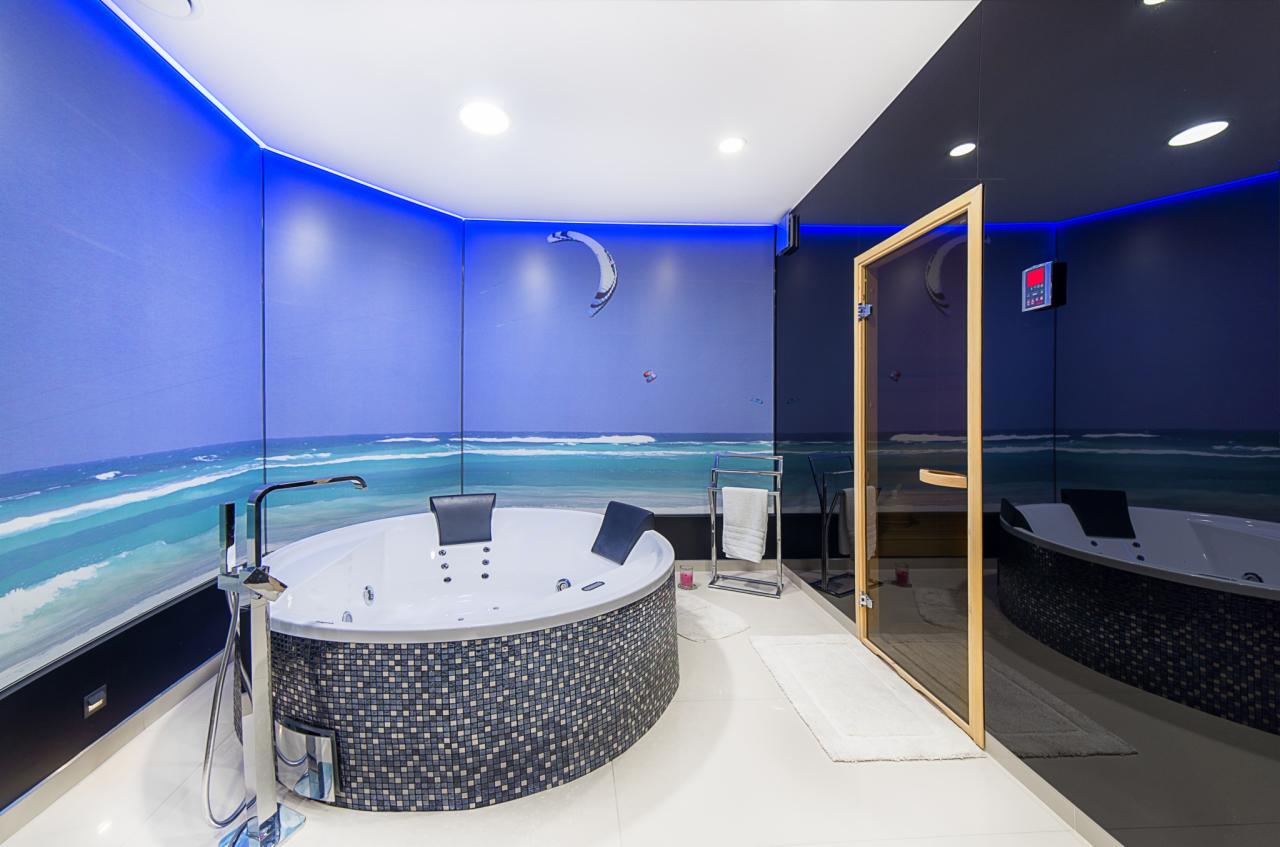 Nowoczesna łazienka z jacuzzi obudowanym mozaiką, fototapetą z motywem morskim i sauną. Oświetlenie LED podkreśla futurystyczny charakter wnętrza.