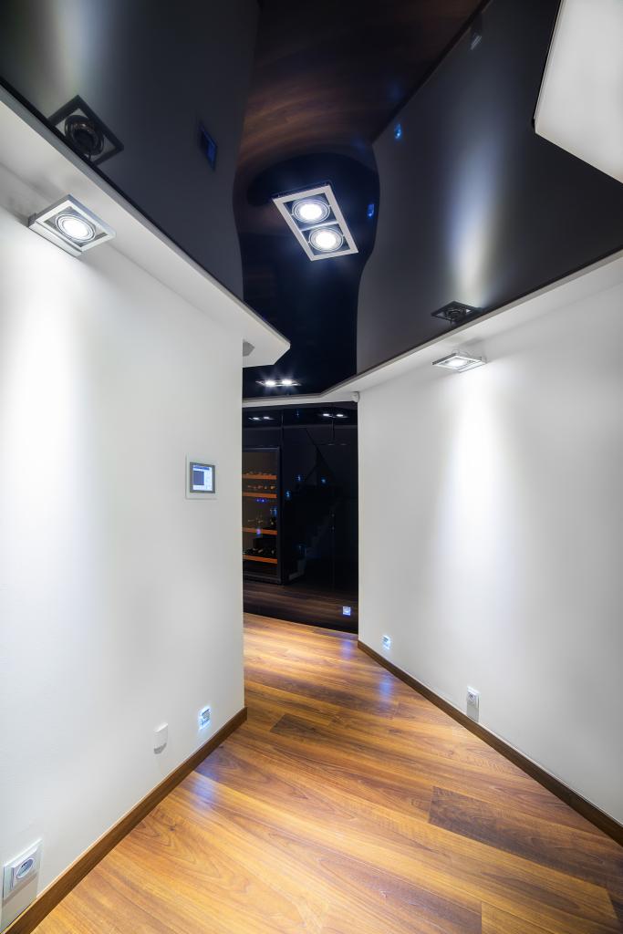 Nowoczesny korytarz z błyszczącym, czarnym sufitem napinanym i punktowym oświetleniem LED. Widoczna drewniana podłoga i minimalistyczne białe ściany.
