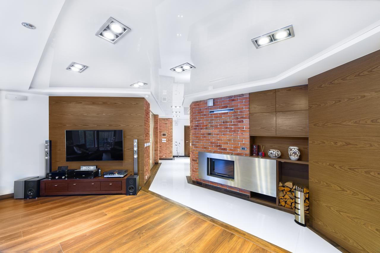 Nowoczesny salon z parkietem, ścianą z drewnianą boazerią i ceglanym kominkiem. Biały, podwieszany sufit z oświetleniem punktowym. Minimalistyczny design.