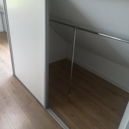 Usługi Stolarskie - Biała szafa z przesuwnymi drzwiami wbudowana w skos poddasza, widoczny metalowy drążek na ubrania i drewniana podłoga.