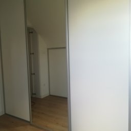 Usługi Stolarskie - Biała szafa przesuwna z lustrem, odbicie wnętrza pokoju z poddaszem i drewnianą podłogą.