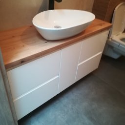 Usługi Stolarskie - Szafka łazienkowa pod umywalkę z blatem drewnianym, wykonana na wymiar w Ryczywole. Białe fronty szafek z delikatnymi frezowaniami, umywalka ceramiczna nablatowa, bateria czarna.