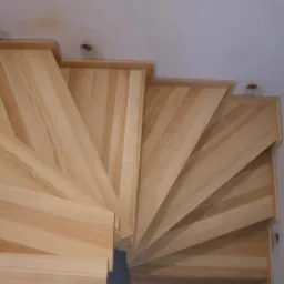 Widok z góry na drewniane, spiralne schody z jasnego drewna, widoczne niedokończone wykończenia sufitu w tle.