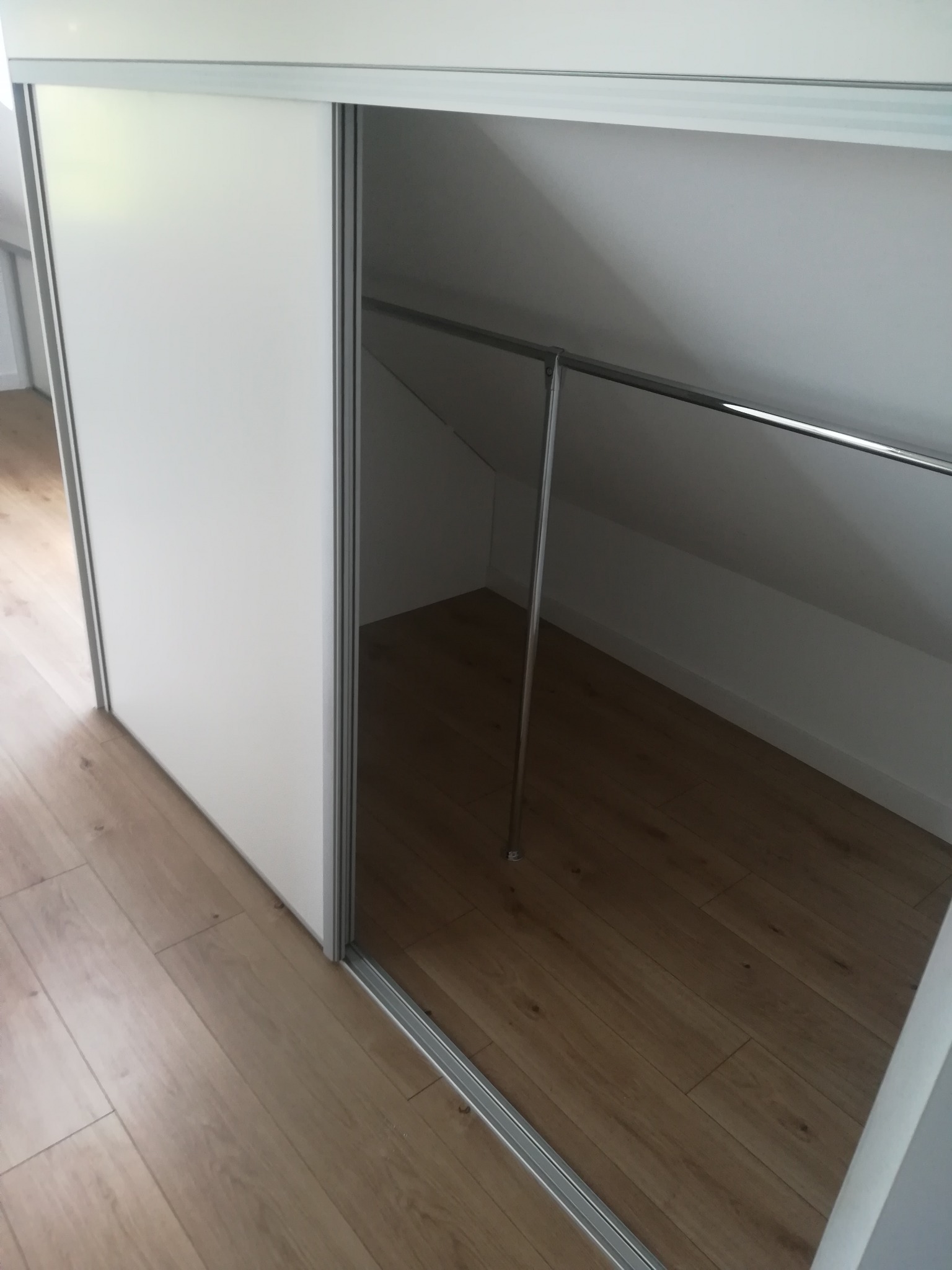 Biała szafa z przesuwnymi drzwiami wbudowana w skos poddasza, widoczny metalowy drążek na ubrania i drewniana podłoga.