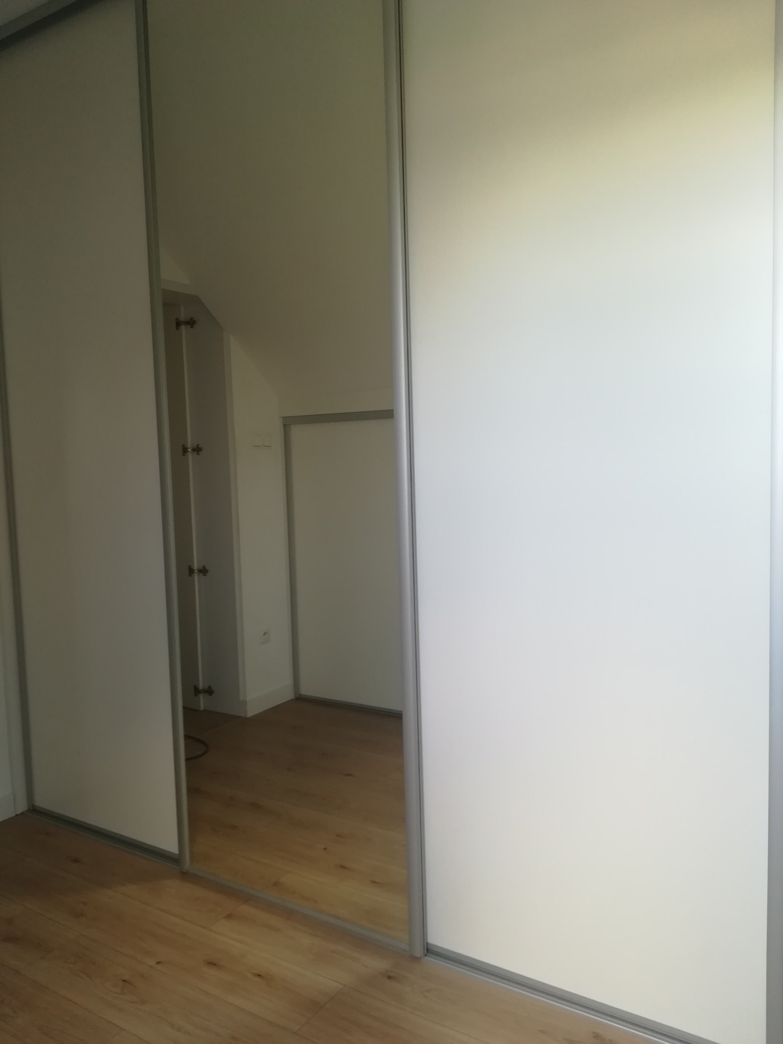 Biała szafa przesuwna z lustrem, odbicie wnętrza pokoju z poddaszem i drewnianą podłogą.