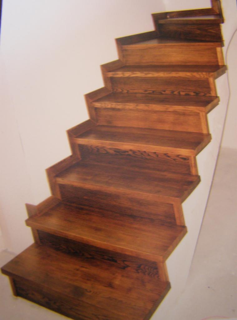 Drewniane schody z ciemnego drewna, wykonane na wymiar w Ryczywole. Stopnie z charakterystycznym wykończeniem, widoczne usłojenie drewna, biała ściana w tle.