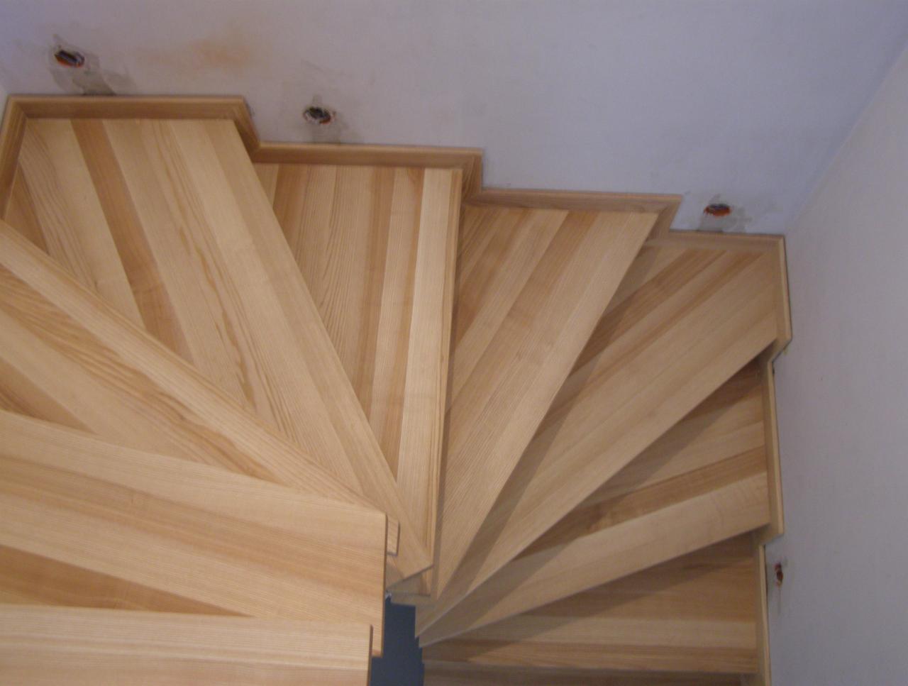 Widok z góry na drewniane, spiralne schody z jasnego drewna, widoczne niedokończone wykończenia sufitu w tle.