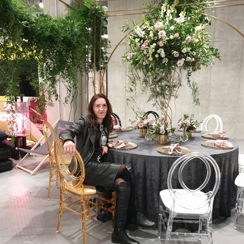 Stylowa aranżacja stołu z kwiatami i dekoracjami, kobieta w skórzanej kurtce siedzi przy złotym krześle, nowoczesne wnętrze z betonową ścianą w tle.