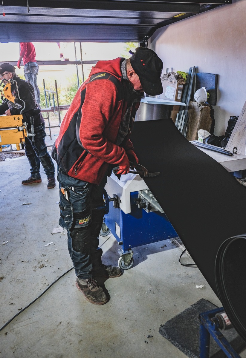 Pracownik w czerwonej bluzie tnie nożycami arkusz czarnej blachy na niebieskim stole warsztatowym na kółkach, w tle inny pracownik przy pracy na rusztowaniu.