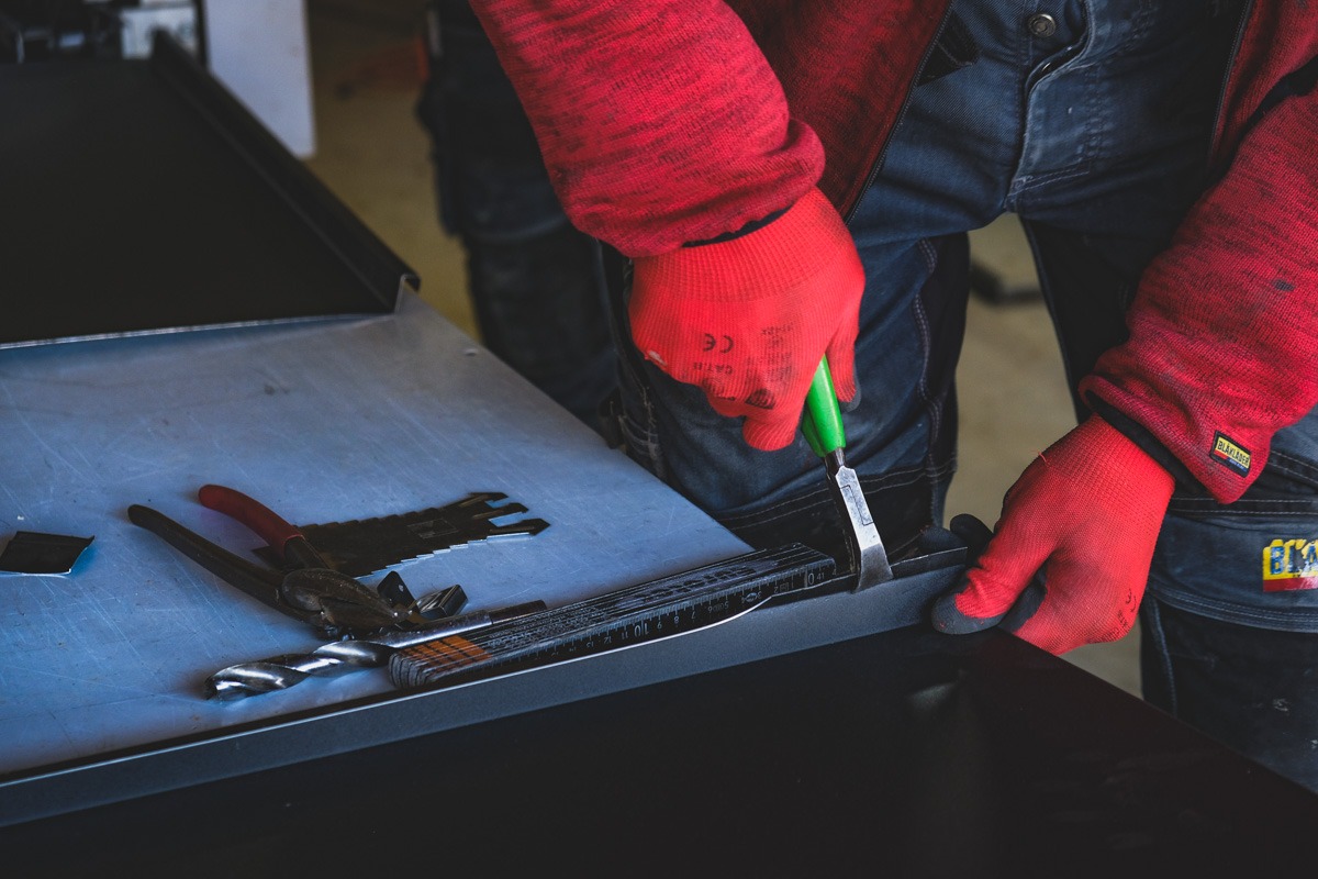 Dekarz w czerwonych rękawicach, używając dłuta z zieloną rączką, obrabia blachę na metalowym stole warsztatowym, obok leżą szczypce, wiertło i miarka.