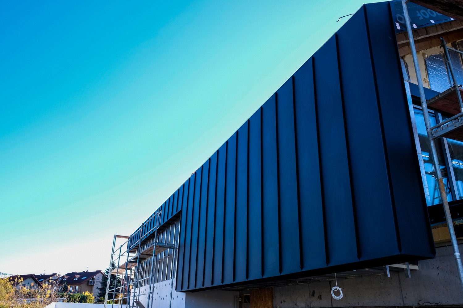 Nowoczesna elewacja budynku w trakcie budowy, pokryta panelami z blachy w kolorze ciemnego granatu, widoczna z dołu na tle jasnego, błękitnego nieba, z fragmentami rusztowania i sąsiednich domów...