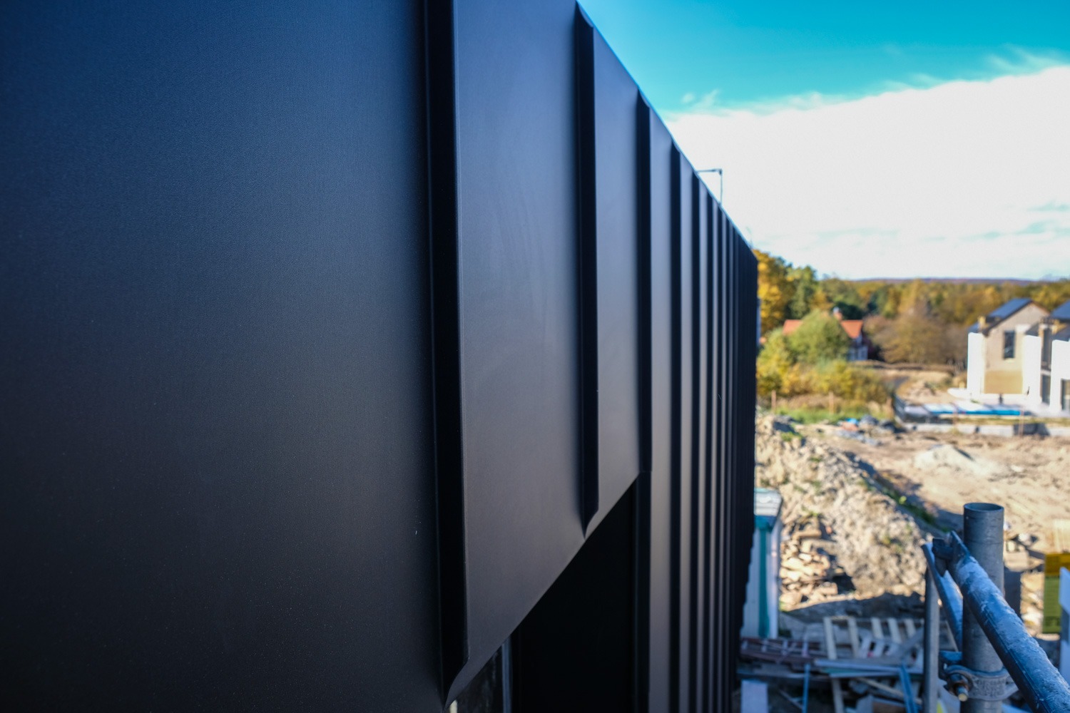 Elewacja budynku wykończona panelami z blachy na rąbek stojący w kolorze antracytowym, widok z rusztowania na tle budowy i jesiennego krajobrazu.