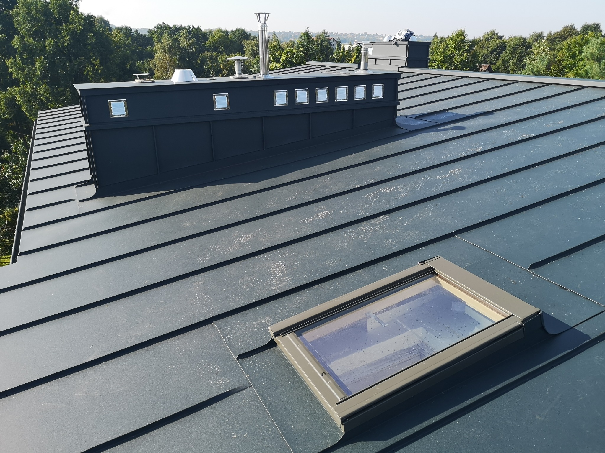Nowoczesny dach z blachy na rąbek stojący w kolorze grafitowym z oknem dachowym i kominem z wbudowanymi luksferami, na tle zieleni drzew.