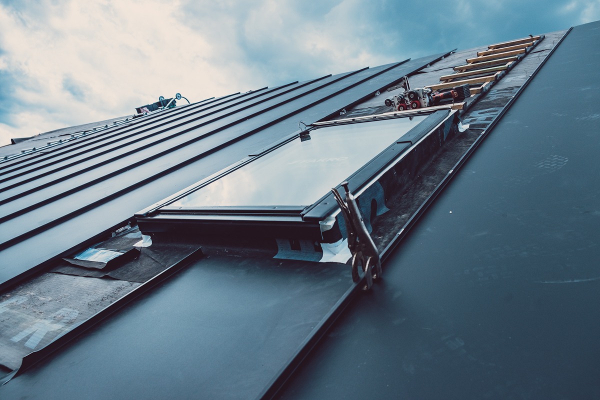 Montaż okna dachowego na metalowym poszyciu z widocznymi narzędziami, fragmentami membrany i drabinką dekarską na dachu w pochmurny dzień.