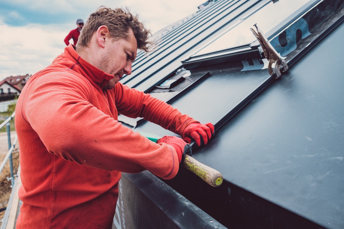 Dekarz w czerwonej bluzie montuje czarne panele dachowe na skośnym dachu, w tle drugi pracownik i zabudowa jednorodzinna.