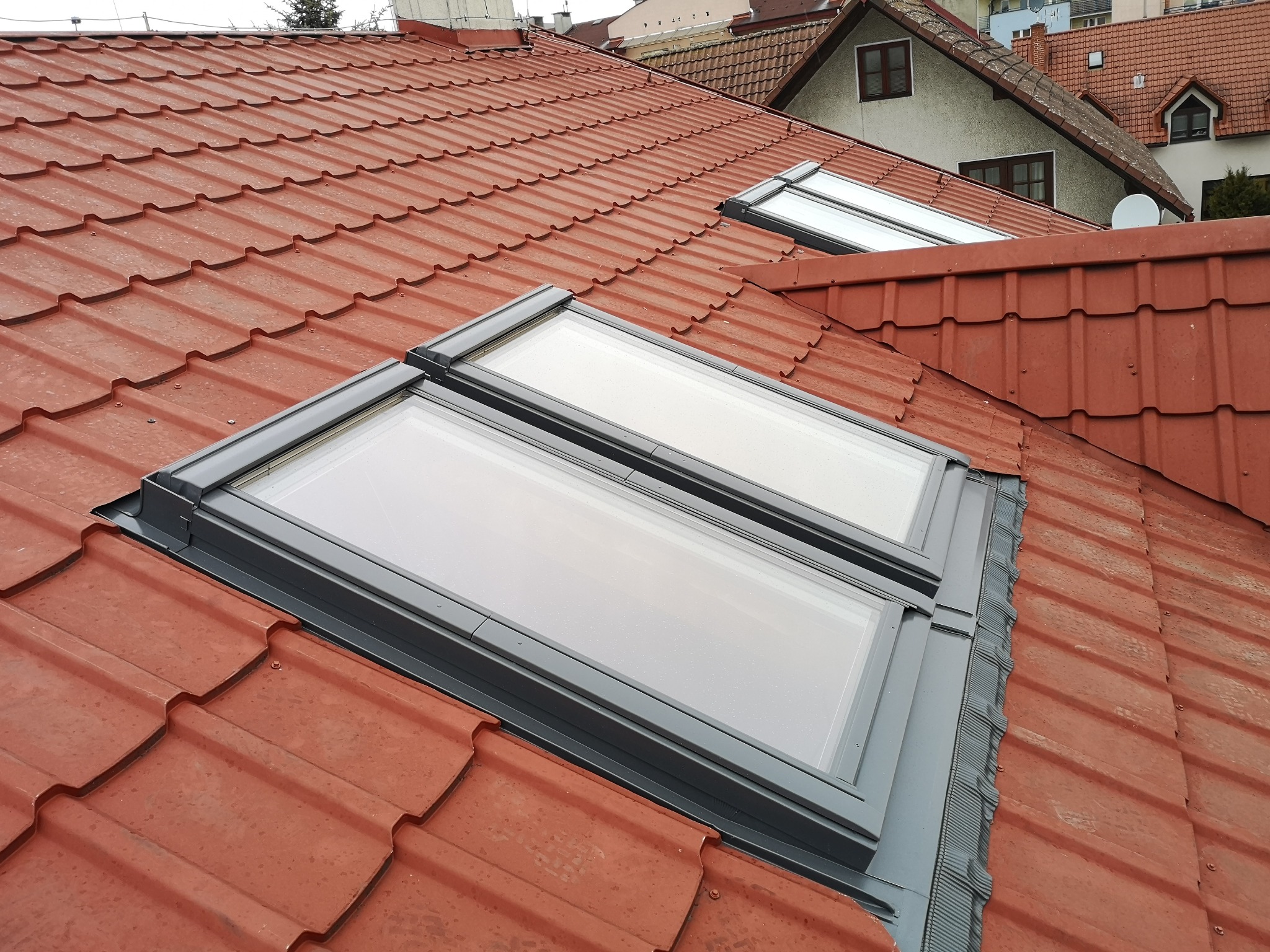 Czerwona dachówka ceramiczna z dwoma oknami dachowymi w szarych ramach, widok z góry na tle zabudowań.