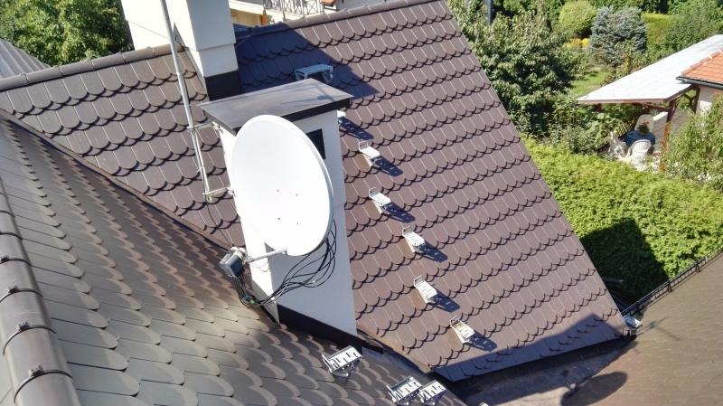 Brązowy dach pokryty dachówką bitumiczną w kształcie łuski, z zamontowaną białą anteną satelitarną i kominem, widoczne stopnie kominiarskie.