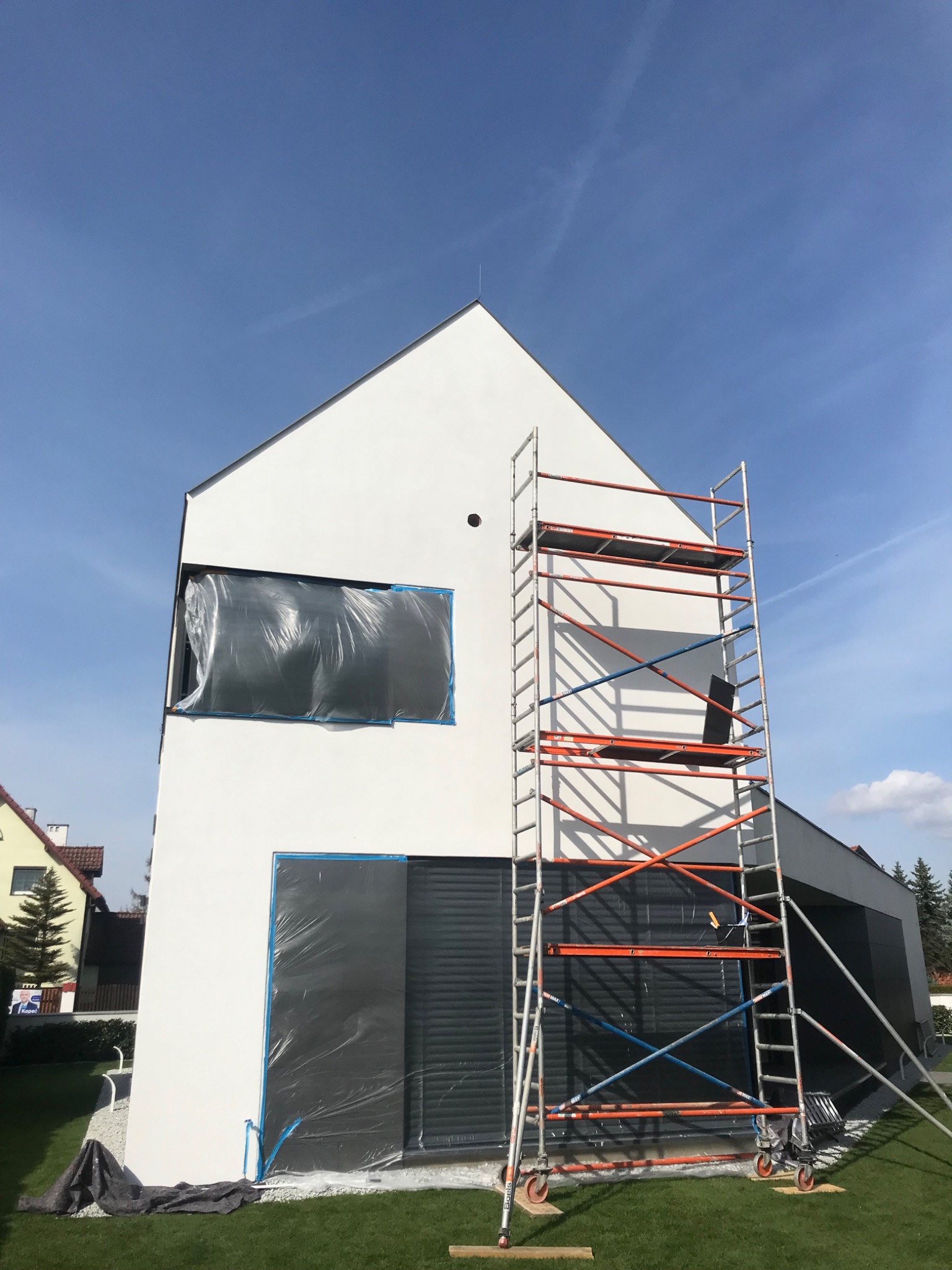 Nowoczesny dom jednorodzinny z białą fasadą w trakcie przygotowań do malowania, okna zabezpieczone folią, rusztowanie ustawione przy ścianie, błękitne niebo w tle.