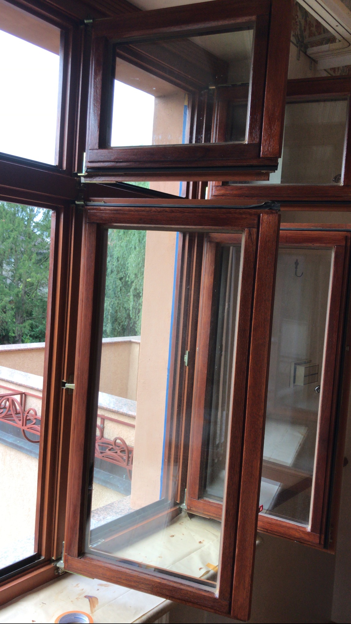 Drewniane okna z szybami, częściowo otwarte, z widocznym balkonem w tle, zabezpieczone taśmą malarską, prawdopodobnie po renowacji lub malowaniu.