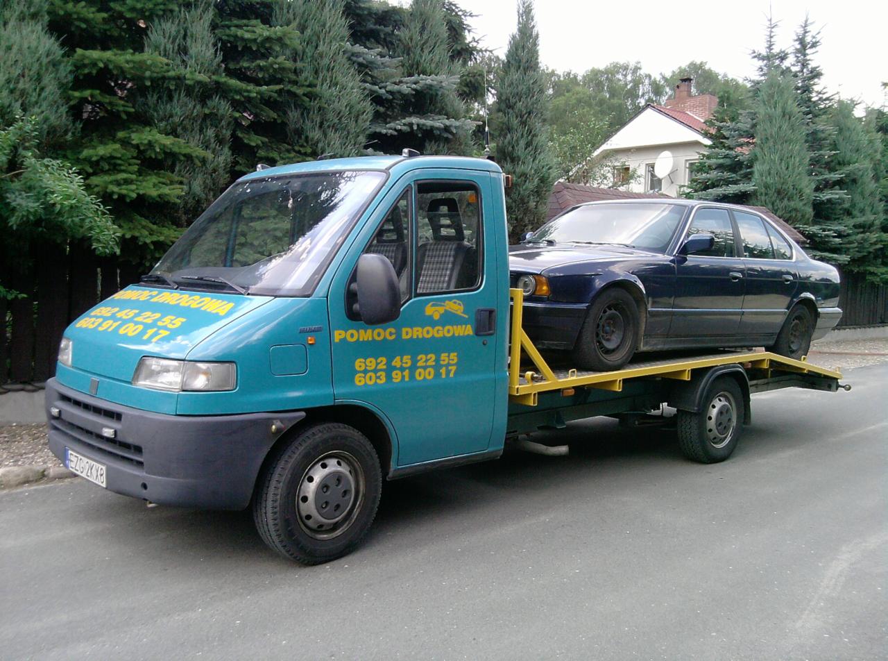 Laweta w kolorze turkusowym z załadowanym granatowym sedanem na tle zieleni i domu. Na lawecie widnieje napis 'Pomoc Drogowa' i numery telefonów.