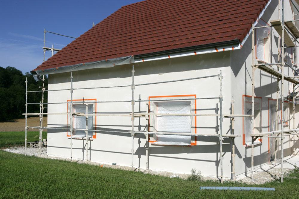 Dom jednorodzinny w trakcie tynkowania elewacji, okna zabezpieczone folią i taśmą, rusztowania dookoła budynku, widoczny fragment dachu z czerwoną dachówką.